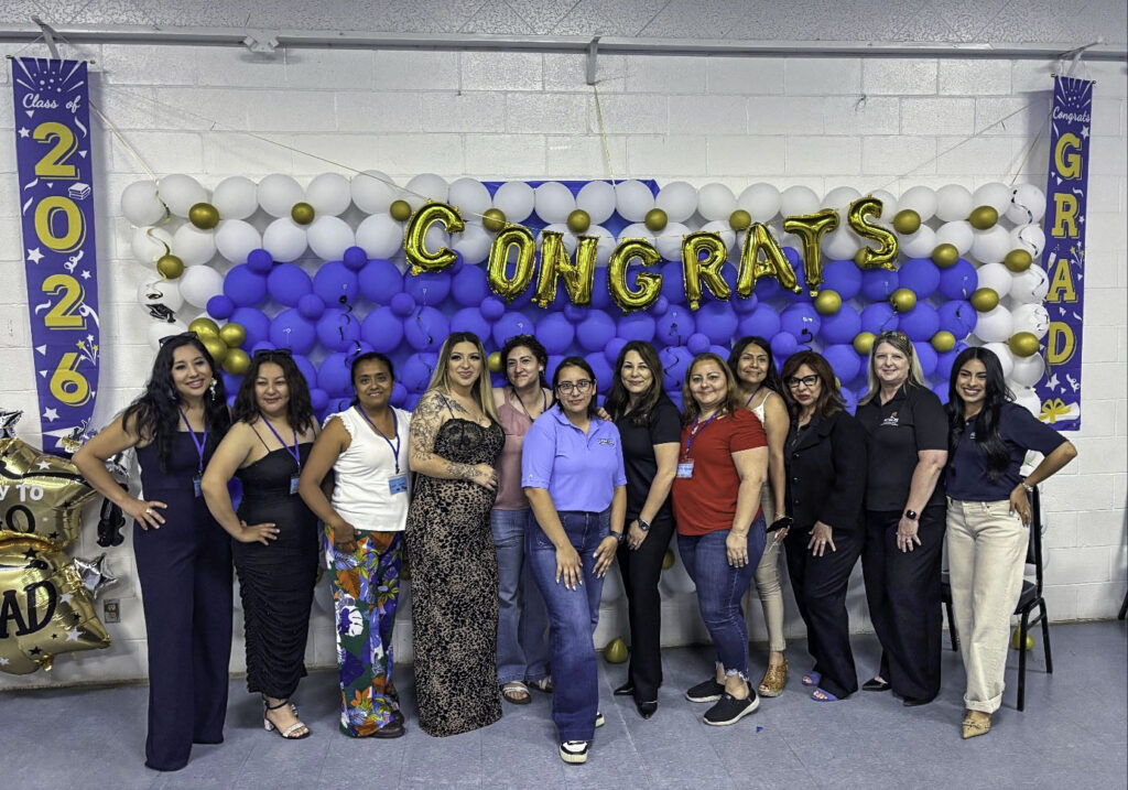 Photo of a group of 12 adult students and staff members from KCSOS stand in a line in front of a balloon wall and a banner reading congrats with two small banners flanking the sides reading "Class of 2026" and "Congrats Grad."