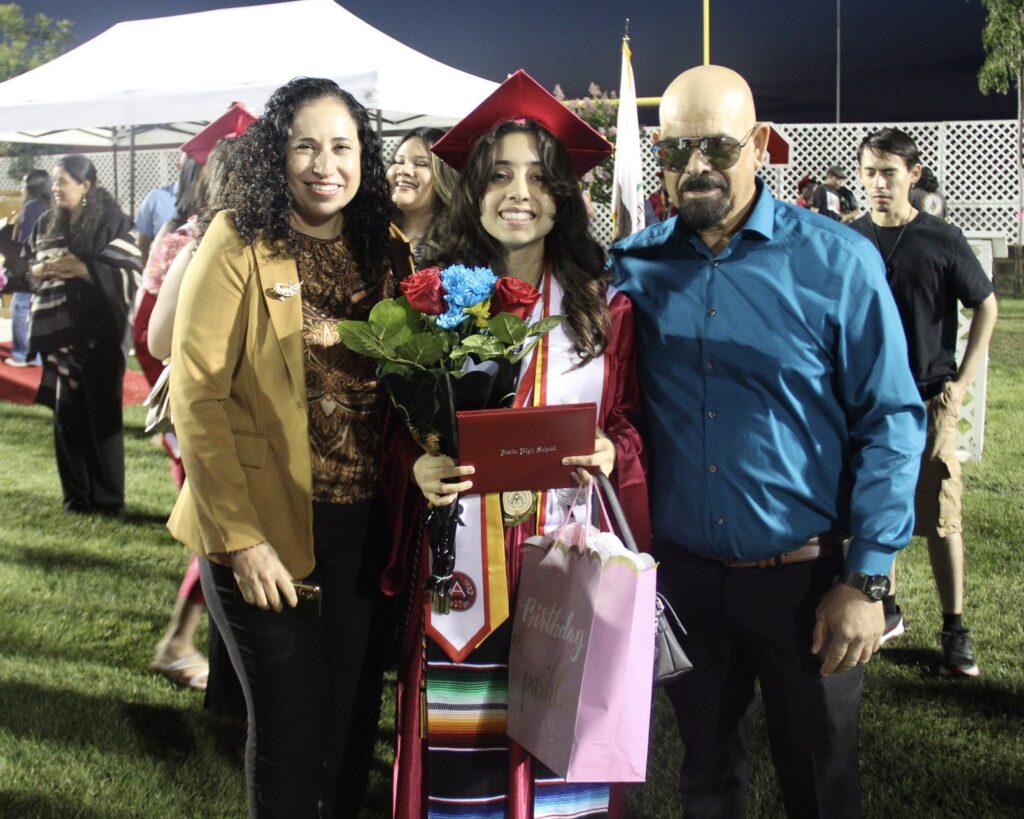 Photo of Acosta Vasquez standing with her parents in her high school regalia.