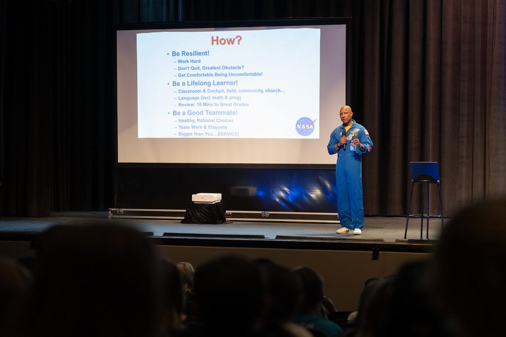 Captain Victor Glover in a blue spacesuit gives a speech on stage, presenting a slide with text on a large screen behind him.
