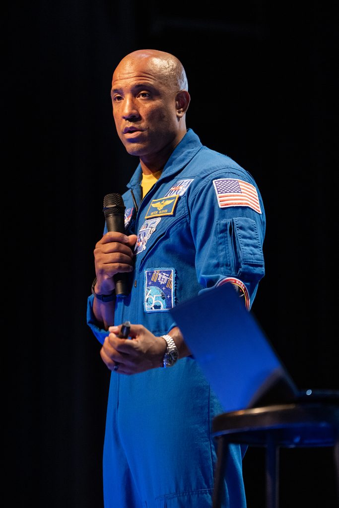 Captain Victor Glover in a blue spacesuit gives a speech on stage.
