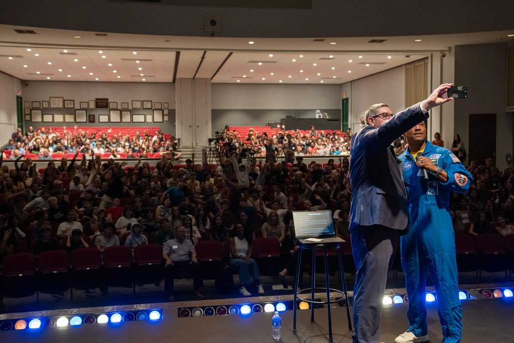 Captain Victor Glover in a blue spacesuit gives a speech to a large audience of students seated in an auditorium.
