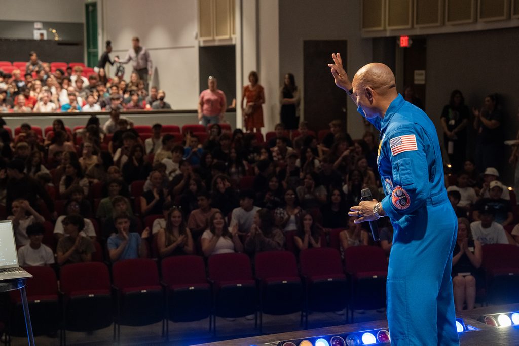 Captain Victor Glover in a blue spacesuit gives a speech to a large audience of students seated in an auditorium.
