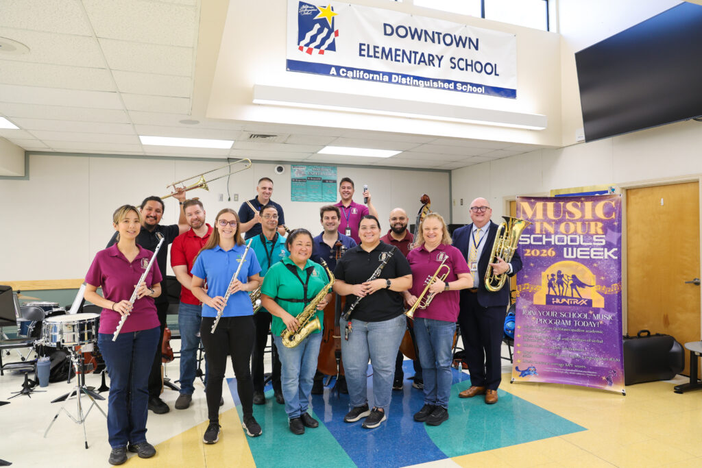 Michael Stone with all district music teachers holding their instruments in front of the Music In Our Schools Poster and Downtown Elementary School banner. 