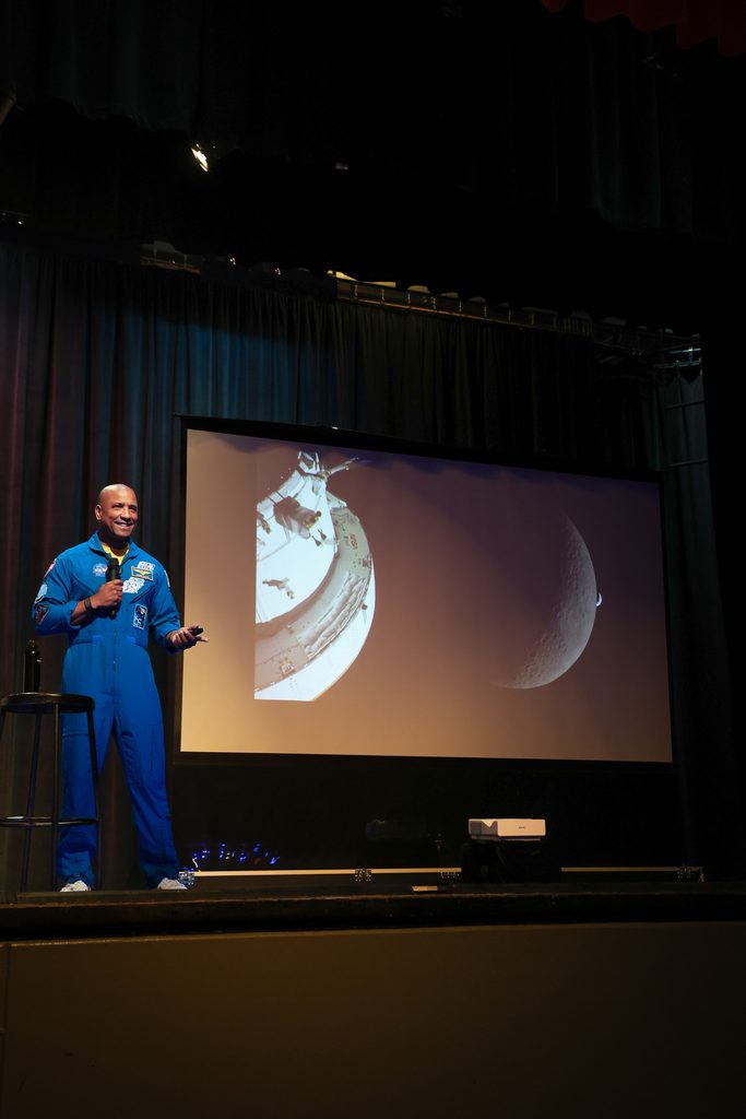 Captain Victor Glover in a blue spacesuit standing on a stage in front of a large projection screen displaying an image of a crescent moon.
